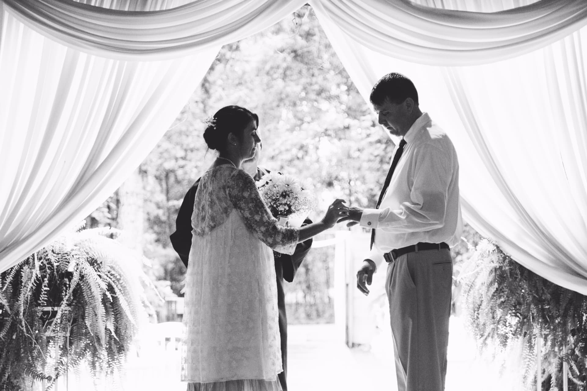 A black-and-white photo of a bride and groom exchanging rings under a draped ceremony canopy