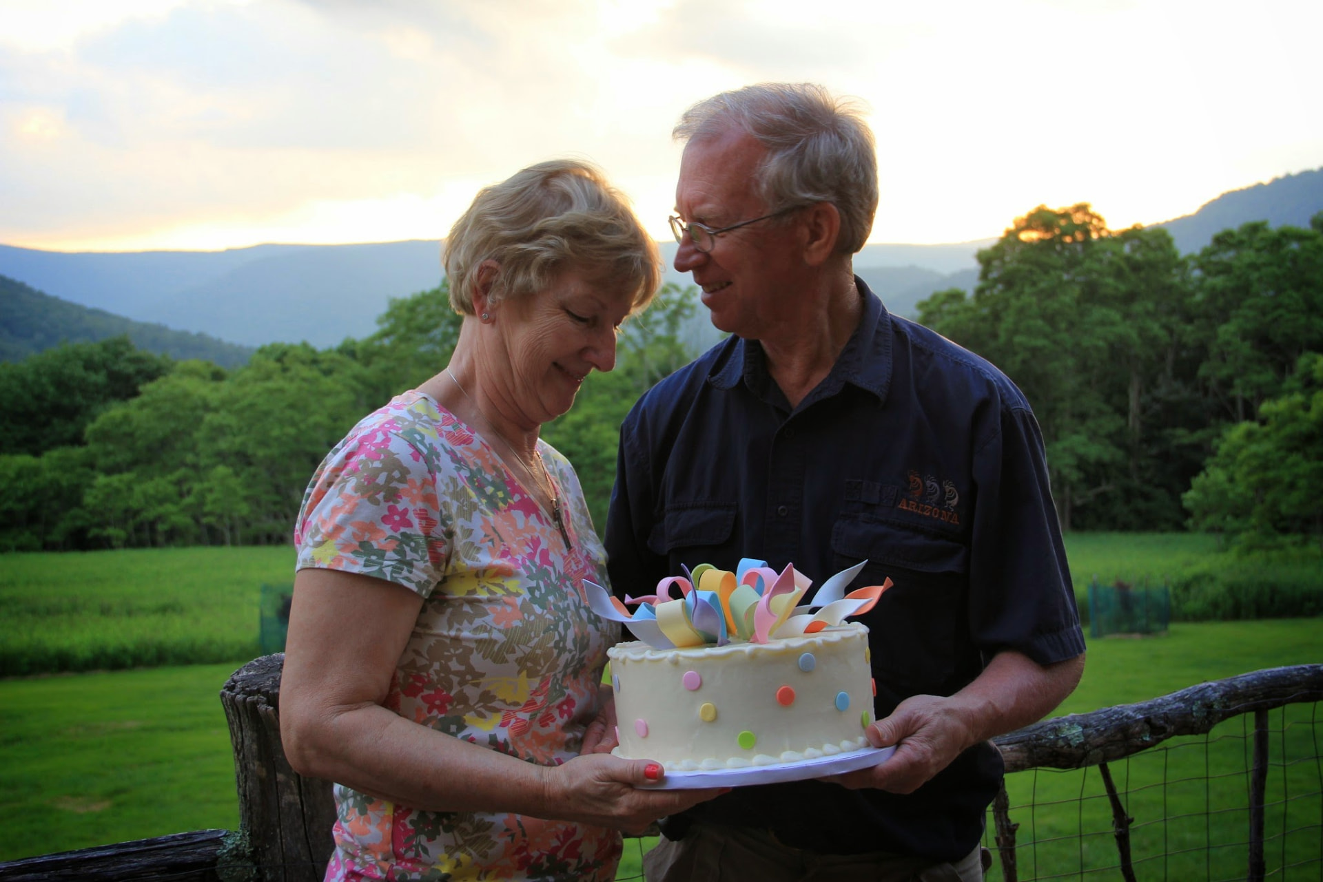 An older couple holding a birthday cake together in front of green mountains