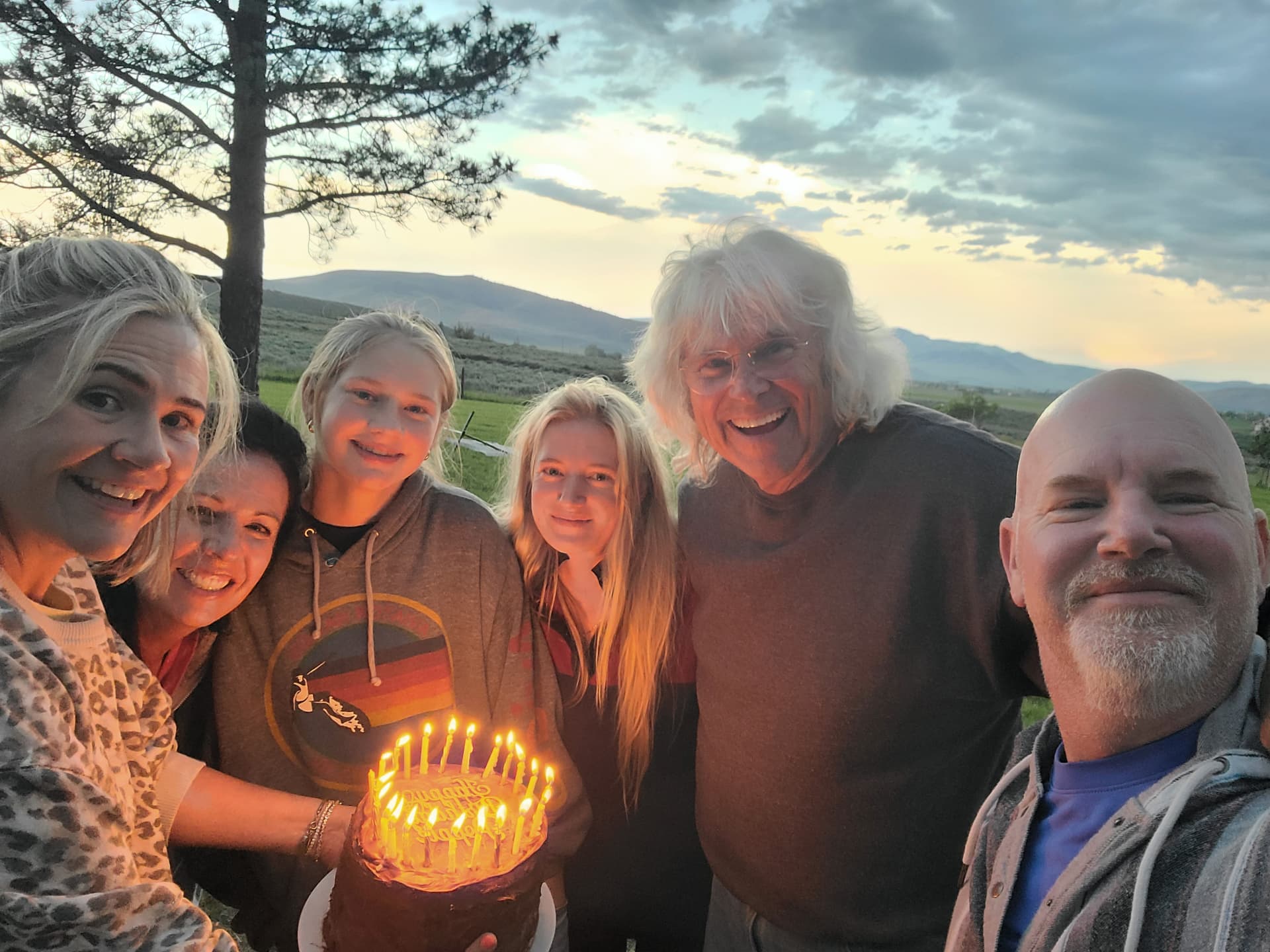 A family crowded together outdoors at sunset around a candlelit birthday cake
